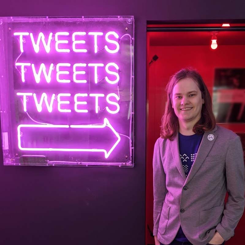 Photo of Jon next to a neon sign saying 'Tweet Tweet Tweet' pointing at them